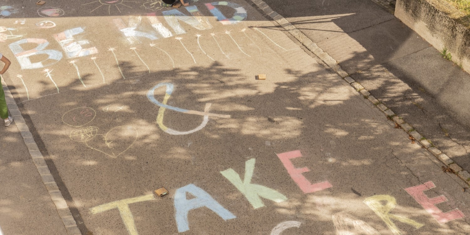 Straßenbemalung "Be Kind & Take Care" mit Kreidefarben. Am Bildrand zwei Kinder mit Kreide in der Hand