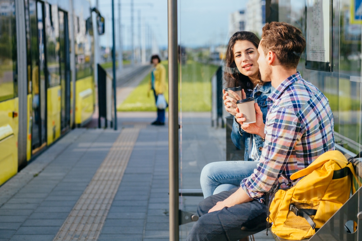 Zwei Personen sitzen bei einer Haltestelle und plaudern, im Hintergrund ein Bus.