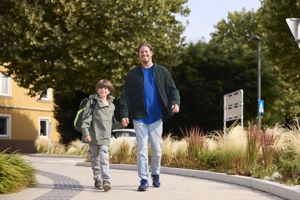 Vater und Sohn gehen zu Fuß auf einem kurvigen Gehweg, im Hintergrund und an den Rändern viel Begrünung. Das Kind trägt einen Rucksack.