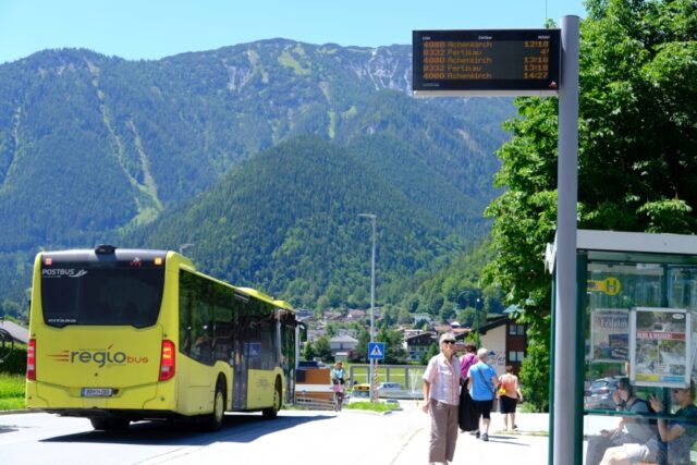 gelber Bus in Tirol an Haltestelle, wartende Menschen. Im HIntergrund ein Berg
