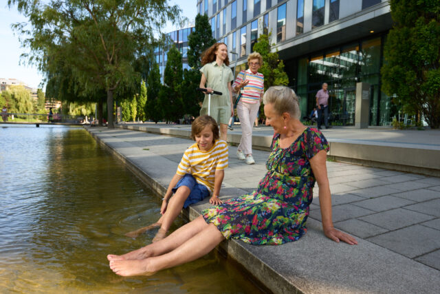Seniorin und Kind sitzen auf einem Betonsteg und halten Füße ins Wasser, im Hintergrund ein Glasgebäude, ein Baum und zwei Personen, die vorbeigehen