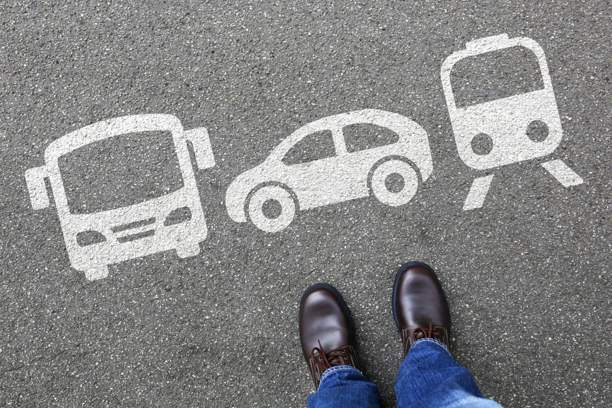 Asphalt mit Piktogrammen eines Busses, eines Autos und eines Zuges, davor sind zwei stehende Beine mit glänzenden Schuhen zu sehen.